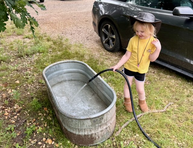 A child in a cowboy hat filling a galvanized water trough with a hose
